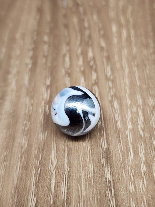 A close-up photograph of a single, round, silver and white swirl-patterned marble resting on a light wooden surface.