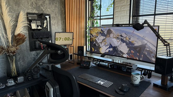 A modern desk setup featuring an ultrawide monitor displaying anime artwork, an ergonomic chair, mechanical keyboard, speakers, and a digital clock reading 07:02.