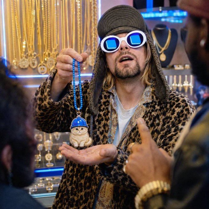 A man wearing a gray cap with ear flaps, white sunglasses, and a leopard-print robe holds up a beaded necklace featuring a jeweled cat pendant wearing a blue cap, standing inside a jewelry shop filled with gold chains and accessories. The image ties to the post's playful promotion of "$BASECAT" season, visually showcasing the bling-covered cat pendant as the centerpiece of the crypto-themed moment.