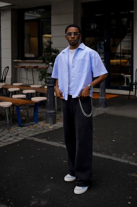 The image features a man wearing a blue shirt and black pants standing outside of a building, posing for a photo. He has his hands in his pockets and appears to be smiling. The surrounding area includes several chairs and dining tables arranged around the scene, suggesting that this might be an outdoor cafe or restaurant setting.