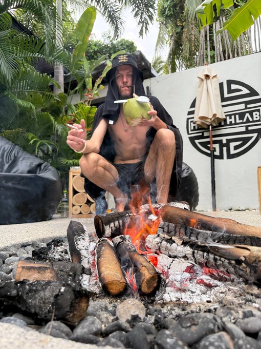 A shirtless man in a hooded robe squats by a fire pit, drinking from a green coconut, with a "HLAB" logo visible on the wall behind him. The tropical, ritualistic setting suggests a laid-back, exclusive vibe fitting the founder of a sophisticated Solana community DAO.