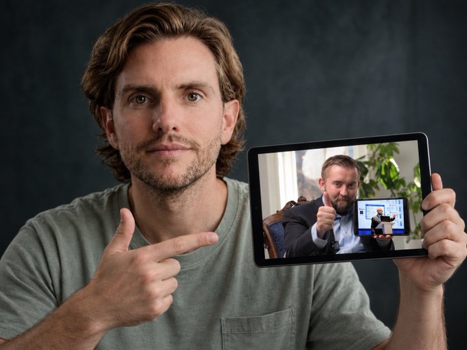 A man points to a tablet screen showing another man pointing to another screen displaying a recursive image of the same pointing men.