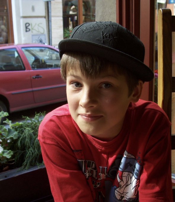 A young boy wearing a black cap with white stitching and a red long-sleeve shirt sits outdoors, with a red car parked on the street behind him.