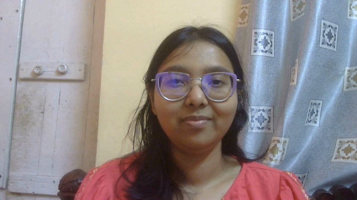 The image features a woman wearing glasses, possibly with red clothing, sitting next to a window. The scene appears to be indoors, as there are no visible outdoor elements or distractions. The focus of the image seems to be on the woman's face and her glasses rather than any other objects in the room.