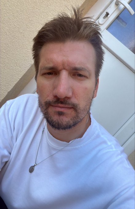 A man with short brown hair and a beard takes a selfie while wearing a white t-shirt and a silver pendant necklace, with a white door and beige wall behind him.