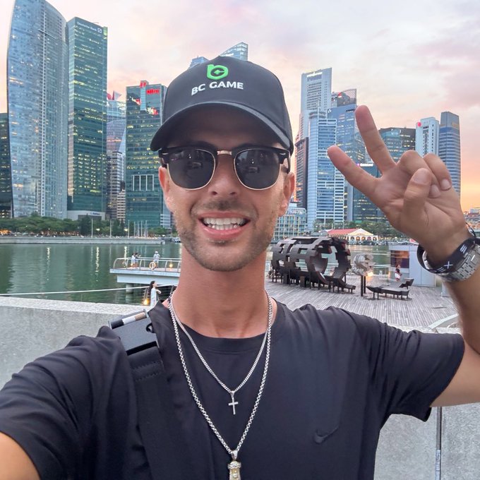 A man wearing a "BC GAME" cap and sunglasses smiles and flashes a peace sign in a selfie, with Singapore's Marina Bay skyline at sunset in the background, visually connecting to the "Gm" greeting and the BC Game collaboration mentioned in the post.
