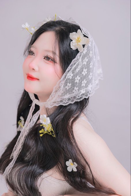 A portrait of a young woman with long dark hair wearing a white lace veil decorated with small flowers and matching floral accessories.