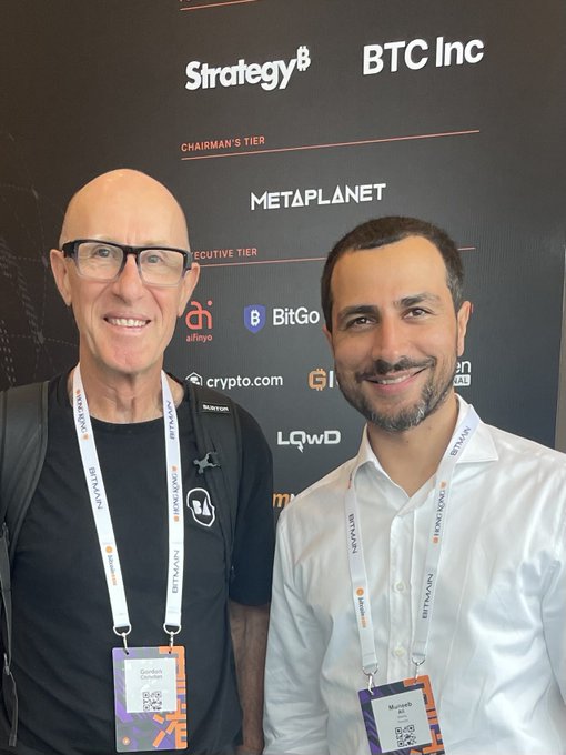 Two attendees wearing "Bitcoin Asia" lanyards pose together in front of a sponsor backdrop featuring BTC Inc, Strategy, Metaplanet, BitGo, crypto.com, and other partner logos. The image appears to capture a meetup with Stacks founder Muneeb Ali at Bitcoin Asia, reflecting the post's tribute to his role in championing Bitcoin as a foundational technology.