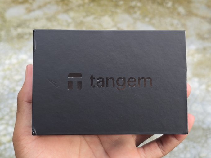 A hand holds up a matte black box with the embossed 'tangem' logo, a brand known for hardware cryptocurrency wallets.