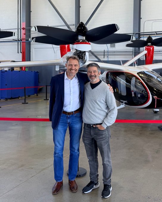 Two men pose smiling in front of a Joby Aviation eVTOL aircraft in a hangar, with the large black propellers and sleek white fuselage prominently displayed behind them. The image relates to the post's playful suggestion that JoeBen Bevirt (Joby's founder) might trade his jetski commute for the company's electric air taxi ($JOBY), while Travis Kalanick appears alongside him as a new investor or partner.