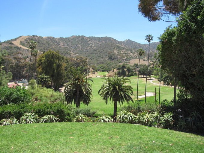 A scenic landscape view of a lush green golf course surrounded by palm trees and dense vegetation, with mountains in the background under a clear blue sky.