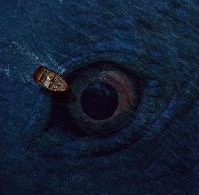 A surreal overhead image of a small wooden rowboat with a single person floating on a dark blue ocean that resembles a giant human eye with a visible iris and pupil.