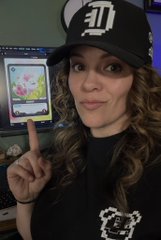 A woman with curly brown hair wears a black t-shirt and a black cap with a white "ET" logo, pointing upward while a computer monitor displays a video game in the background.