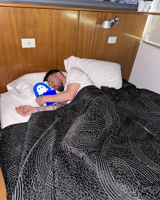 A bearded man sleeps peacefully in bed, cuddling a blue pillow with a white ghost-like character design, wrapped in a black patterned comforment in a wood-paneled room. The image humorously pairs with the caption "1 eth and a dream," as the single ether cryptocurrency is represented by the lone "1" alongside the dreaming sleeper clinging to his crypto-themed plush.