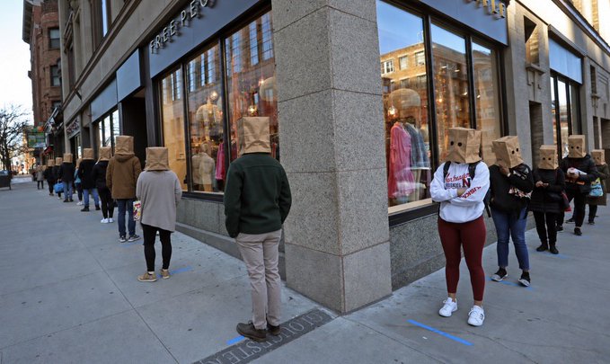 A line of people standing on a city sidewalk wearing brown paper bags over their heads, spaced apart in front of a Free People clothing storefront.