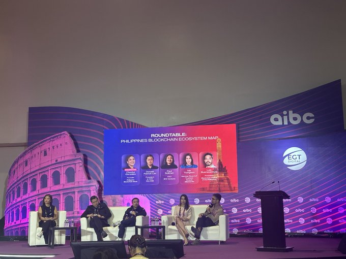 The image shows a group of people sitting around a table, with a large screen behind them displaying information about blockchain adoption at the AIBC World conference. The speakers are identified as @helloluis and @heypaulroots, who are discussing the topic on stage. There is also a microphone visible in front of one of the speakers.