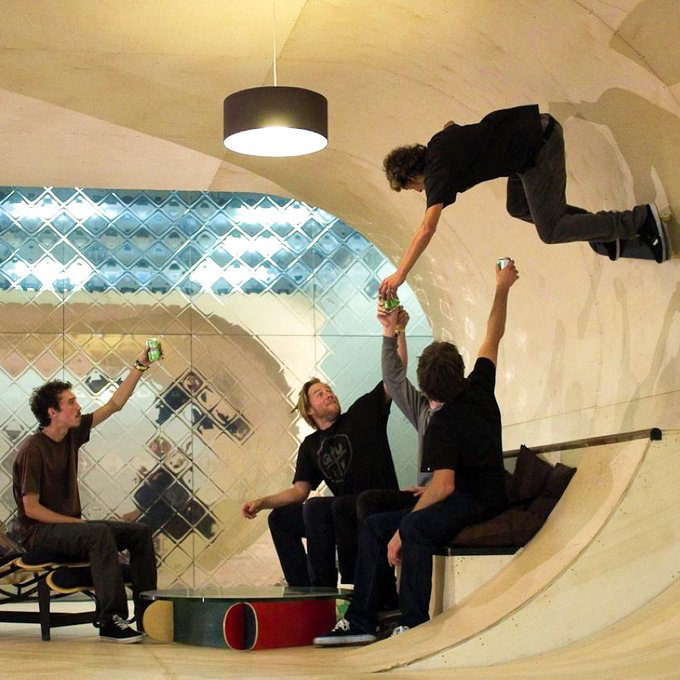 Four people are gathered in a uniquely designed room with curved walls and mirrored tiles, with one person appearing to defy gravity on the ceiling while holding a can with the others below.