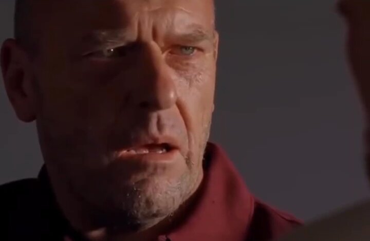 The image shows an older man with a beard wearing a red shirt, looking at someone who appears to be holding a gun. The man's expression suggests that he is upset or angry about something related to the gun. The person holding the gun seems to be making a threat against the man, and it appears as if they are in a tense situation.
