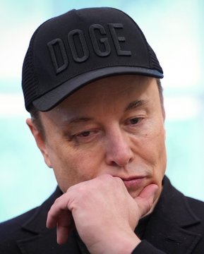 A man wearing a black cap with 'DOGE' written on it, resting his chin on his hand in a contemplative pose.