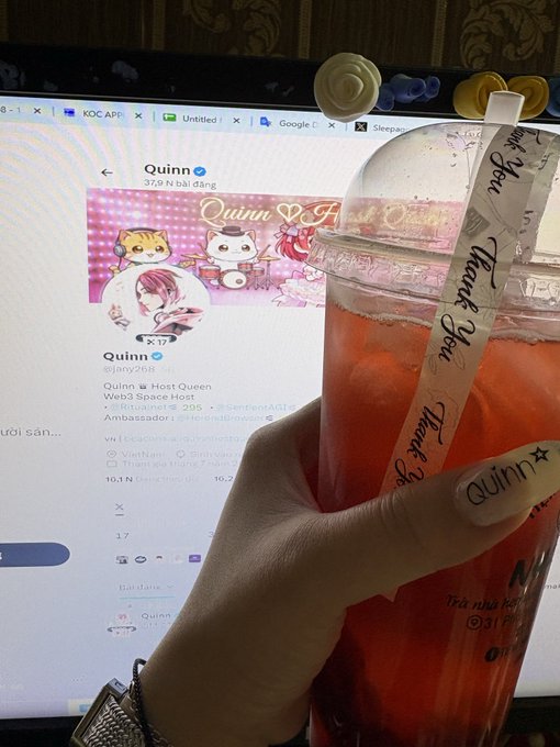 A hand holds up a plastic cup of vibrant red iced tea, with a straw wrapper reading "Thank You" and "Quinn," while a computer screen in the background displays the Zalo profile of "Quinn" with 16.2K followers and the handle "@qinn26B," echoing the post's celebratory "Hello new week!" vibe with its bold splash of red.