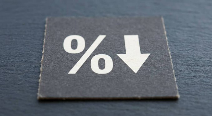 A close-up photograph of a dark gray square label or tag on a wooden surface, printed with a white percent sign (%) and a downward-pointing arrow, symbolizing a decrease or reduction.