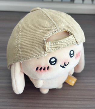 A cute plush toy with a smiling face and pink blush cheeks wears a beige baseball cap backwards, sitting on a wooden surface.