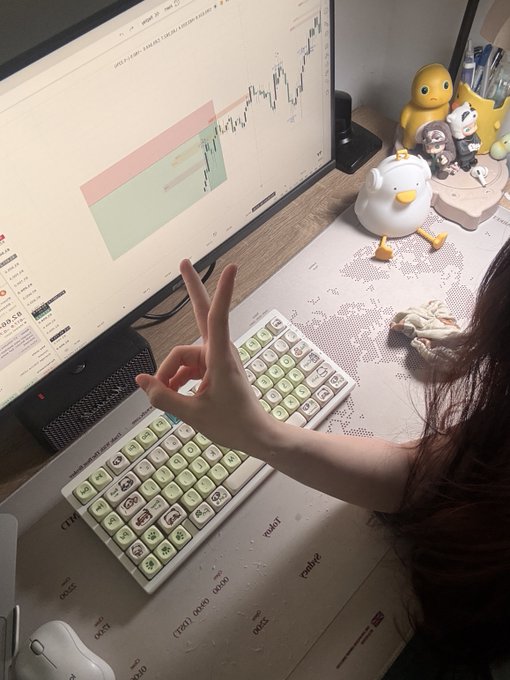 Over-the-shoulder view of a person making an 'OK' hand gesture while sitting at a desk with two mechanical keyboards, looking at a monitor displaying trading charts, with rubber duck figurines nearby.