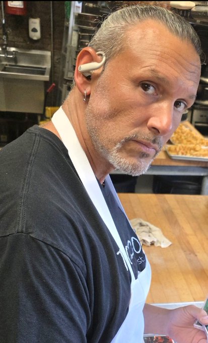 A man with gray hair and beard, wearing an earpiece and a white apron over a black shirt, in what appears to be a kitchen setting.