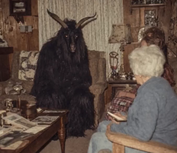 A person in a black goat costume with horns sits in a chair, facing two elderly women in a wood-paneled living room setting.