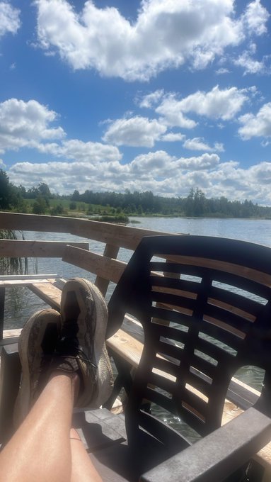A person relaxes on a deck with their feet propped up in worn running shoes, gazing out at a serene lake under a stunning sky filled with painterly white clouds against blue. The peaceful scene contrasts with the post's celebration of stock gains ($HIVE, $BMNR, and $SPCX up 5%), capturing the tranquil "great way to start the week" moment the author enjoyed while overlooking the water.