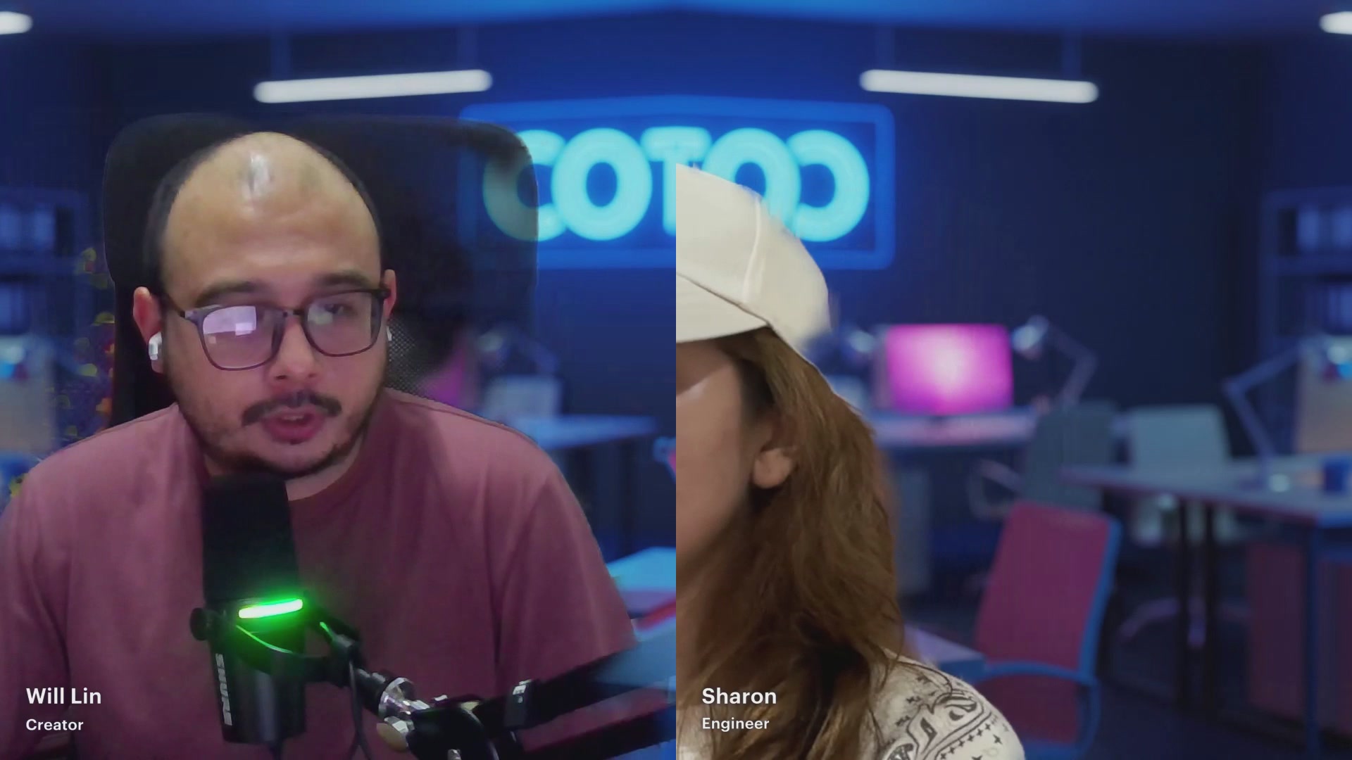 The image shows a man wearing glasses sitting at a desk with a microphone, while another person wearing a hat is standing behind him. They are engaged in an online chat or video call using a messaging app like WhatsApp or Messenger. The background of the image features a blue wall and a pink chair, creating a casual atmosphere for their conversation.