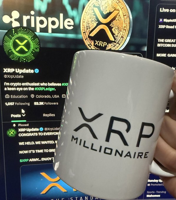 White coffee mug with 'XRP MILLIONAIRE' text held in front of a screen showing Ripple/XRP branding and an XRP Update social media profile (@XrpUdate) with a pinned post about #XRPLedger.