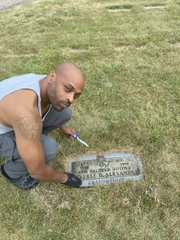 A man crouches beside a grave marker for Geraldine Masterson, displaying a newspaper that reads "BABE'S BIGGEST FAN'S TRIBUTE" as a tribute to his late mother, honoring her memory 25 years after her passing from Multiple Sclerosis.