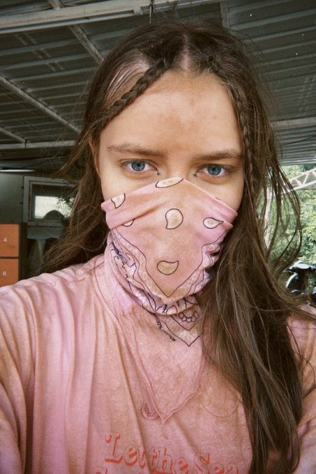 A close-up selfie of a young woman with braided hair wearing a pink bandana face covering and a pink t-shirt that reads 'Let the Sea'.