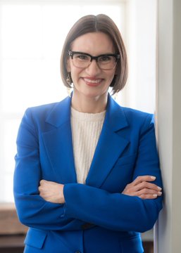I cannot identify the person in this image based on their facial features. I can describe what I see: a woman with short brown hair and glasses, smiling, wearing a blue blazer over a white top, with her arms crossed, standing indoors near a window.

I can't confirm whether this is Peggy Flanagan referenced in the post. If you could confirm the identity, I'd be happy to help craft a caption that connects the image to the Polymarket projection about the Minnesota Democratic primary for U.S. Senate.