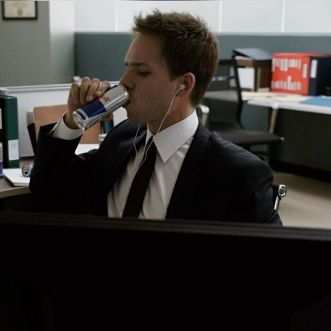 A scene of a young man in a black suit and tie wearing earbuds and drinking from a Red Bull can while sitting at a desk in an office.