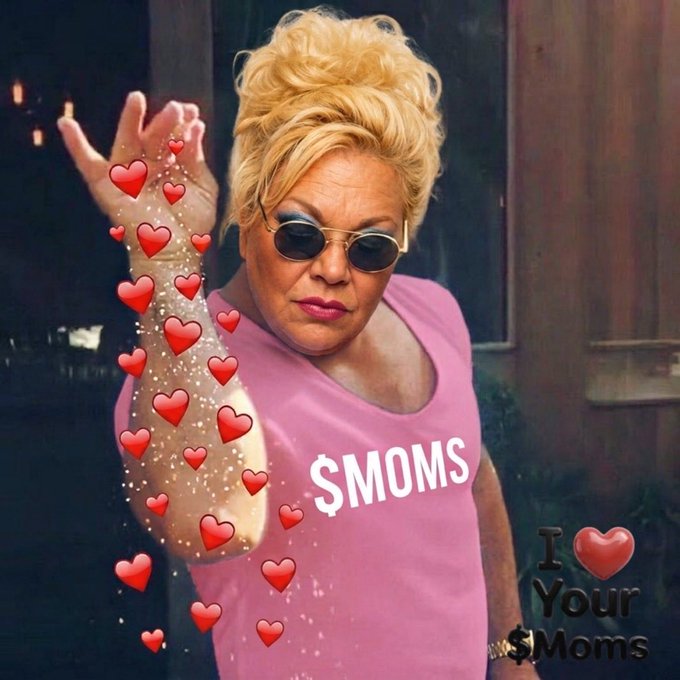 A woman wearing sunglasses and a pink "$MOMS" t-shirt poses with her arm raised as animated heart emojis cascade around her, while an "I ❤ Your $Moms" graphic appears in the bottom corner—tying directly into the meme-style promotion for the "$MOMS" token.