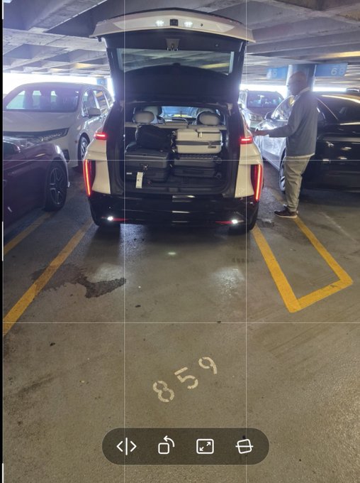 A view from inside a vehicle showing the open rear hatch of an SUV (BMW iX) loaded with luggage in a parking garage, with a person standing beside it.