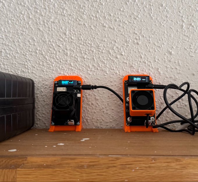 Two orange Miboxer-brand battery chargers with digital displays sit on a wooden floor against a textured white wall, with a black case nearby.