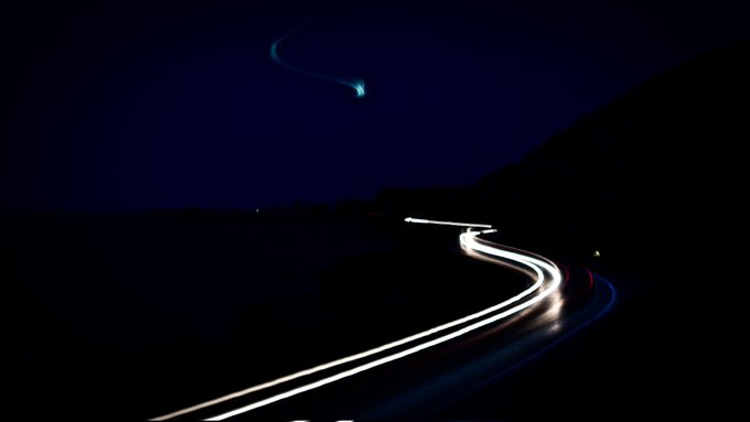 The image shows a long exposure photograph of a winding road at night, with white car lights streaking across the scene as they travel along the curve of the road. The sky above appears dark, creating an atmospheric effect that enhances the sense of motion in the photo. The image is posted on SOVRREIGN gallery and is described as a "Good Morning dear friends Available 1/1 long exposure photography on SOVRREIGN gallery on Objkt curated by @KOLLECTOR_OG Edt: 1/1 Price: 170 XTZ".