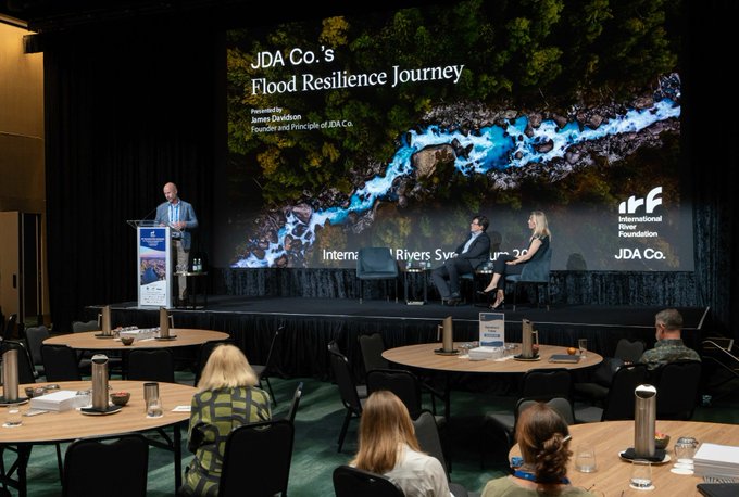 A presentation titled 'JDA Co.'s Flood Resilience Journey' is shown at an IRF (International River Foundation) event, with a speaker at a podium and panelists seated on stage.