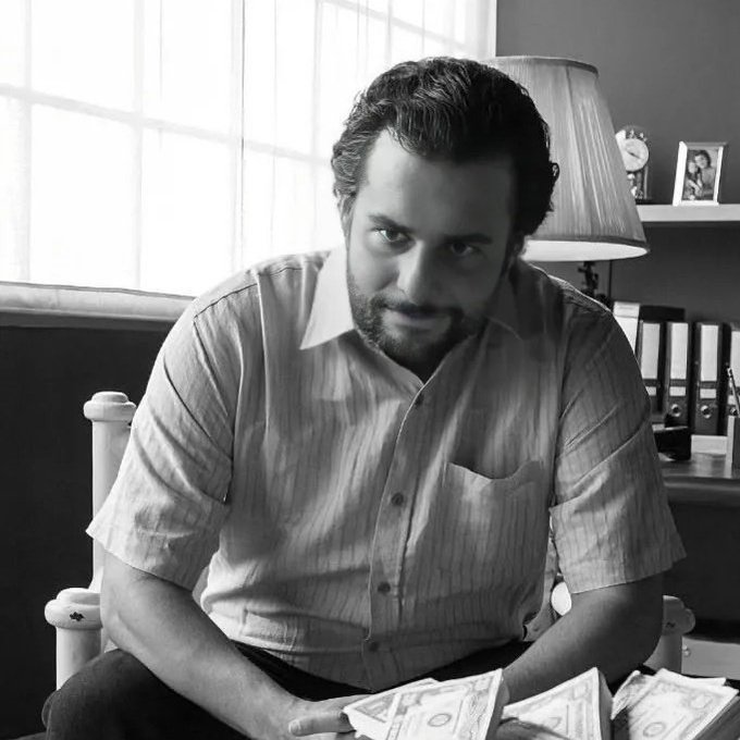 A black-and-white photo shows a bearded man in a short-sleeved button-up shirt sitting at a desk with stacks of cash in the foreground.