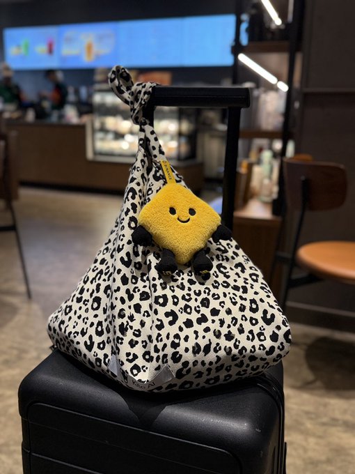 A leopard-print drawstring bag with a smiling yellow plush charm sits on a black suitcase handle in what appears to be a station concourse.