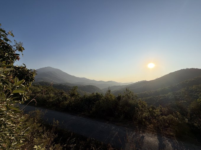 A scenic landscape photo taken at sunrise or sunset, showing rolling forested mountains under a clear blue sky with the sun glowing brightly on the right side.
