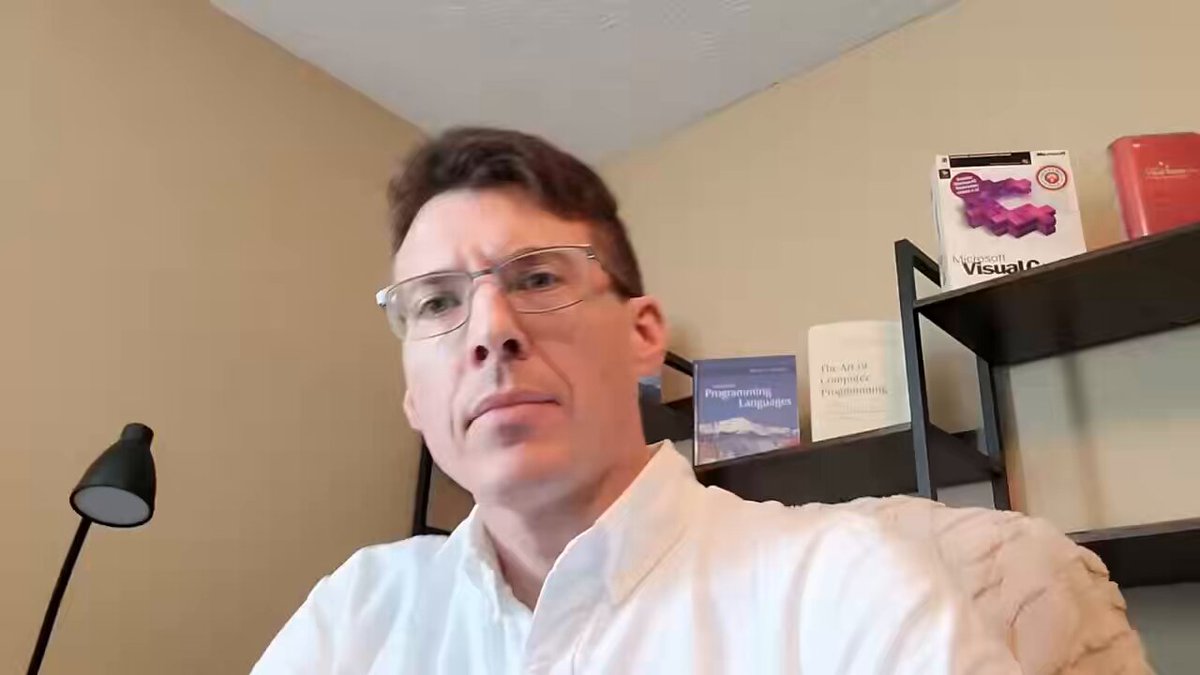 The image shows a man wearing glasses sitting at a desk with a lamp next to him. He has a serious expression as he looks directly into the camera, possibly participating in an online meeting or webinar. The background features several books on a shelf, suggesting that this could be a workspace or study area for the man.