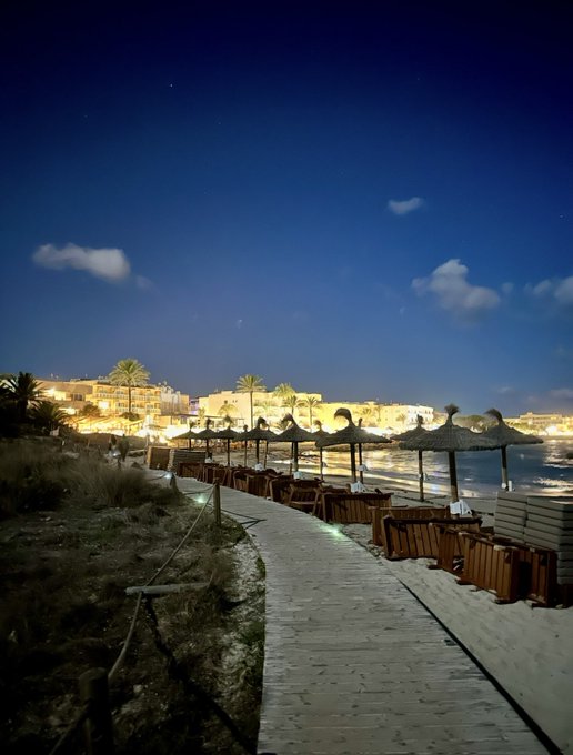 A serene nighttime beach scene featuring a wooden boardwalk lined with thatched umbrellas and lounge chairs, illuminated by soft lights with a starry sky and distant resort buildings in the background. The peaceful coastal setting perfectly matches the "good night" message, evoking a tranquil end-of-day moment at a seaside destination.