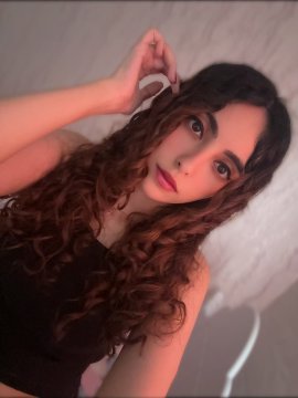 A young woman with long, curly auburn hair takes a selfie while lying down, wearing a black top, with one hand raised near her head against a white background.