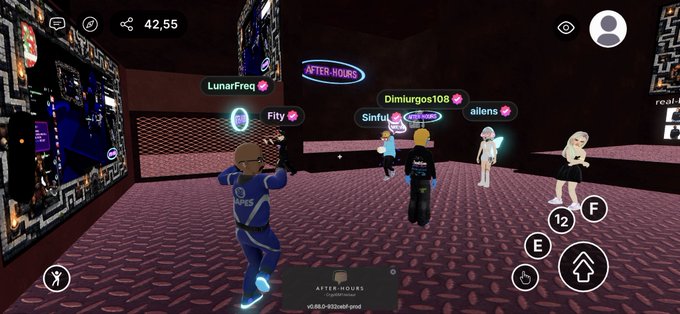 The image shows a group of people playing a video game together, specifically in a virtual world called @decentraland. The players are wearing blue suits and holding weapons as they engage in combat within the game. There are at least five visible characters in the scene, with one player standing out from the rest due to their unique attire - a blue suit with neon lights on it. The group is gathered around a large screen that displays the game's interface, which includes various options and controls for the players to interact with.