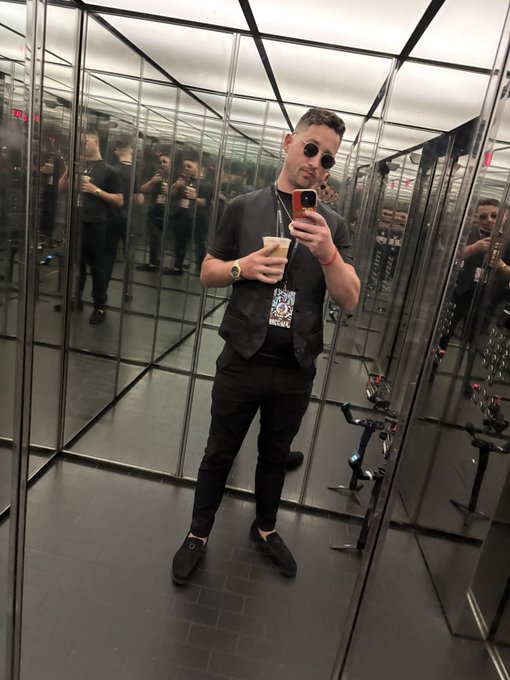A man in all-black attire and sunglasses takes a mirror selfie in a mirrored room, holding a phone and a coffee cup.