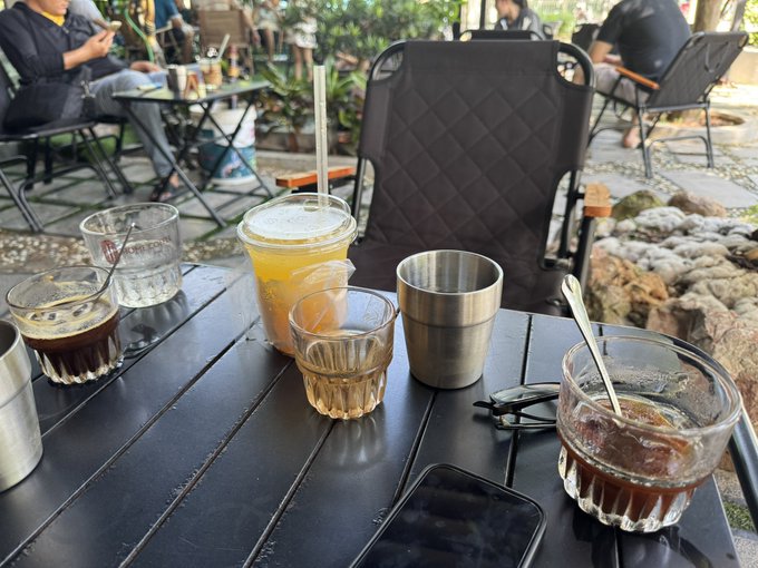 An outdoor cafe scene with multiple drinks including iced coffee, orange juice in a plastic cup, and water on a dark wooden table, with empty folding chairs and people in the background.